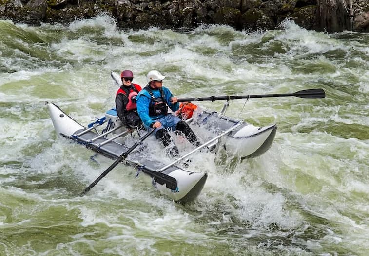Two rafters paddling in the turmoil river stream in a two floating sides boat. On the background a rocky shore hit by the stream waves 